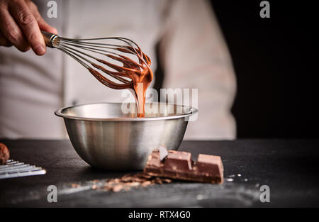 Lo Chef Sbattere il cioccolato fuso in un acciaio inossidabile terrina usando un vecchio vintage frusta in un close up sulla sua mano Foto Stock