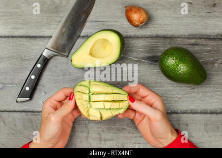 Vista del piano portapaziente, donna mani di avocado con tagli, preparazione, rimozione di polpa. Complesso di avocado, semi e il coltello grigio sulla scrivania di legno accanto ad essa. Foto Stock