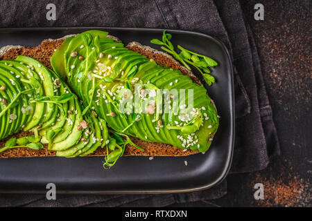 Avocado toast con pane di segale e semi su una piastra nera, sfondo scuro. Foto Stock