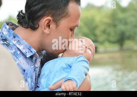 Padre a piedi attraverso il parco vicino al lago con tenendo la sua todler mentre baciare il suo Figlio bambino Foto Stock