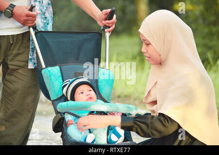 Musulmani asiatici hijabi il padre e la madre a piedi attraverso il parco con il figlio in passeggino Foto Stock