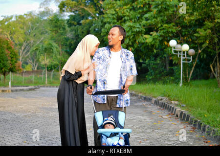 Musulmani asiatici hijabi il padre e la madre a piedi attraverso il parco con il figlio in passeggino Foto Stock