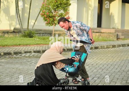 Musulmani asiatici hijabi il padre e la madre a piedi attraverso il parco con il figlio in passeggino Foto Stock