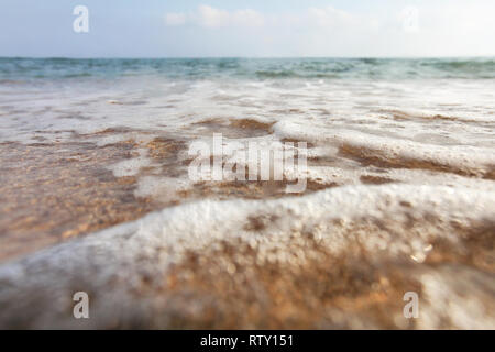 Angolo basso, fotocamera sul terreno, la lente coperta con acqua scende a sottolineare wetness - close up di mare poco profondo lavaggio onde di sabbia sulla spiaggia. Abstract marin Foto Stock