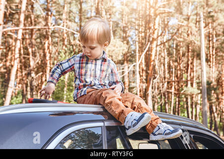 Ritratto di un bambino seduto sulla cima di un auto nera e giocare con il suo giocattolo. Forest in background Foto Stock