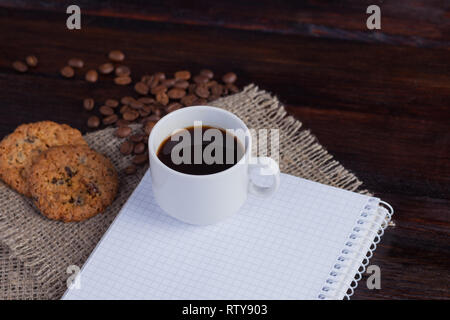 White tazza di caffè con caffè in chicchi vicino e biscotti e blocco note in una gabbia per i record su tessuto di lino sul vintage scuro dello sfondo. Foto Stock