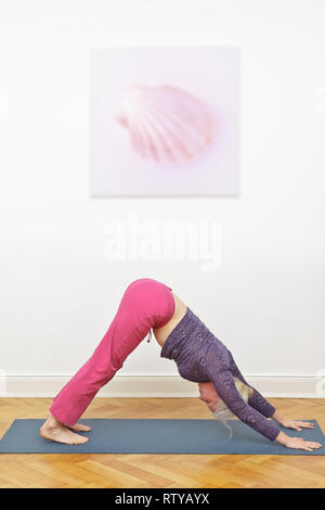 Senior donna esercizio di yoga su un tappetino a casa, asana cane verso il basso, shell immagine sul muro bianco in background Foto Stock