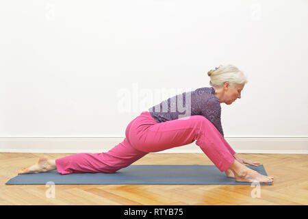 Senior donna con i capelli grigi facendo esercizi yoga a casa davanti ad un muro bianco, posizione dragon intro Foto Stock