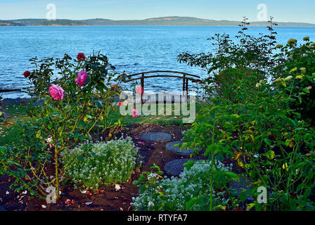 Un giardino con rose affacciato sul bordo dell'acqua e un piccolo ponte pedonale presso la Locanda del Mare vacation resort nei pressi di Nanaimo sull'Isola di Vancouver Foto Stock