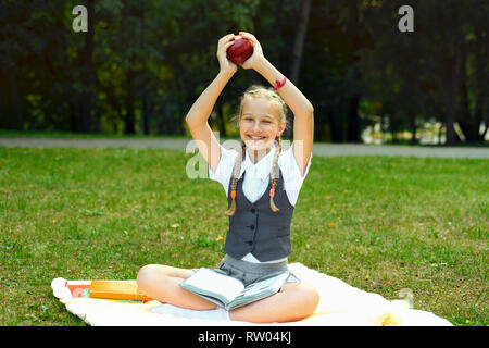 Studente in uniforme è sorridente e felicemente sta tenendo in mano una mela rossa overhead. schoolgirl seduta su una coperta in un parco con libri e facendo home Foto Stock