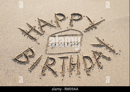 Buon compleanno messaggio scritto a mano liscia in sabbia con una fetta di torta di pianura sulla riva di una spiaggia vuota Foto Stock