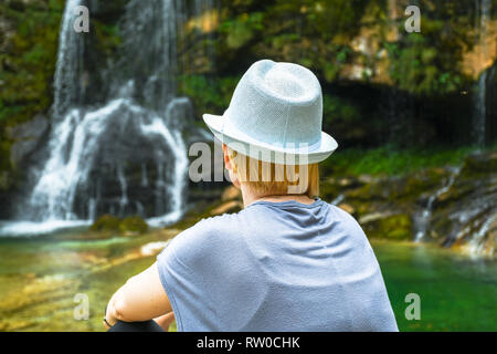 Donna con capelli biondi e white hat cercando, seduta rilassante e dal laghetto naturale e cascata in background Foto Stock