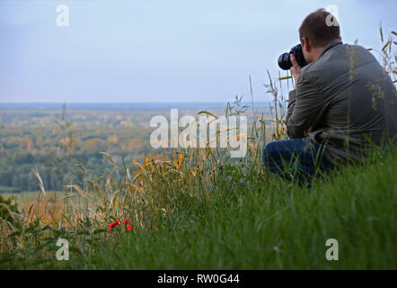 L'uomo con la fotocamera si siede su un alto colle verde accanto alle orecchie di frumento e papavero rosso fiori Foto Stock