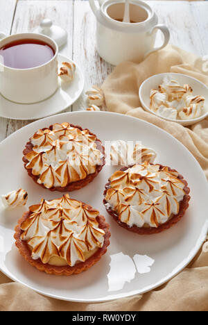 Close-up di Lemon Meringue Pie mini rosolato con picchi di meringa servito con tè, mini marshmallows e marrone zucchero organico su un tavolo di legno con luce Foto Stock
