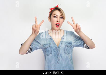 Ritratto di funny bella giovane donna in casuale blu denim shirt con il trucco e la fascia rossa in piedi con la vittoria o segno di pace e guardando a camma Foto Stock