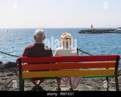 Vista posteriore di una coppia di anziani seduti su un multi-colore banco, godendosi un po' di sole invernale all'estero & guardando al mare; Lanzarote, Febbraio 2019 Foto Stock