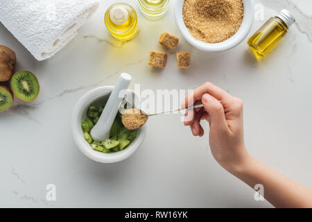 Vista ritagliata della donna aggiungendo lo zucchero di canna nel pounder con kiwi sulla superficie bianca Foto Stock