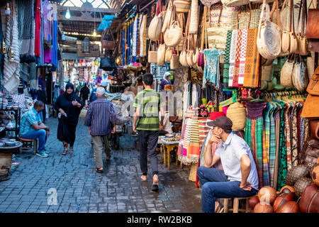 Merchants sedersi di fronte al loro store-front come gli amanti dello shopping a piedi e navigare sulla stradina nel quartiere della medina di Marrekech, Marocco Foto Stock