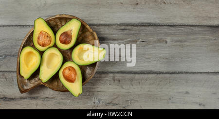 Vista dall'alto in basso, Avocadi tagliati a metà, seme visibile posto sul vecchio scolpita in legno ciotola e grigio scrivania di legno. Spazio per il testo sulla destra. Foto Stock