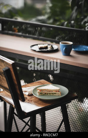 Abstract Scena di prima colazione pasto impostare compresi tonno sandwich, hot lungo nero caffè e brownie cheese cake su legno outdoor bar. Fine settimana mattina acti Foto Stock