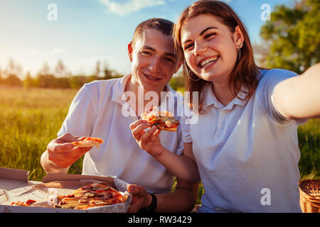 Coppia giovane mangiare la pizza fuori e tenendo selfie nel parco. Donna e uomo avente picnic al tramonto. Ragazzi divertirsi e godere il cibo. Foto Stock