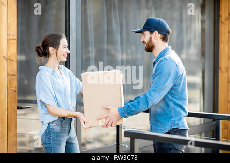 Uomo di consegna portando merci confezionate ad una giovane donna in piedi client insieme all'aperto di fronte alla casa moderna. Foto Stock