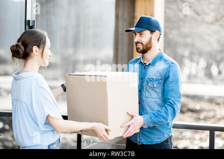 Uomo di consegna portando merci confezionate ad una giovane donna in piedi client insieme all'aperto di fronte alla casa moderna. Foto Stock