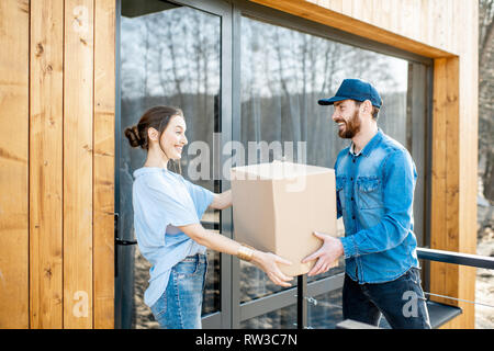 Uomo di consegna portando merci confezionate ad una giovane donna in piedi client insieme all'aperto di fronte alla casa moderna. Foto Stock