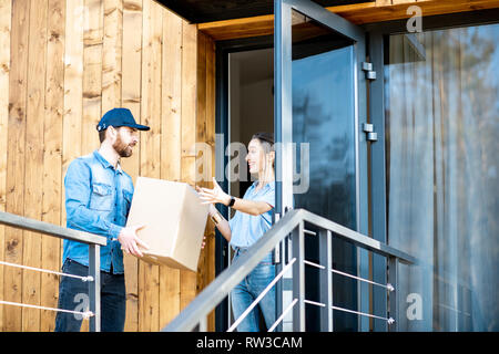 Uomo di consegna portando merci confezionate ad una giovane donna in piedi client insieme all'aperto di fronte alla casa moderna. Foto Stock