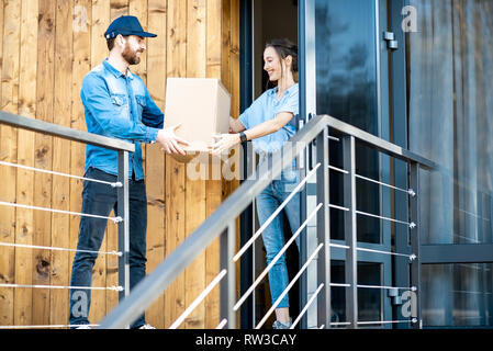 Uomo di consegna portando merci confezionate ad una giovane donna in piedi client insieme all'aperto di fronte alla casa moderna. Foto Stock