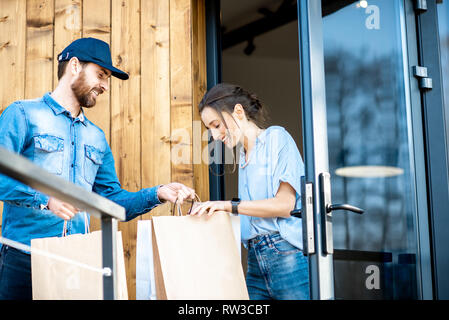 Uomo di consegna portando un po' di merci imballate in sacchi di carta per una giovane donna client a casa. Acquistare vestiti online e concetto di consegna Foto Stock