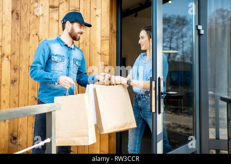 Uomo di consegna portando un po' di merci imballate in sacchi di carta per una giovane donna client a casa. Acquistare vestiti online e concetto di consegna Foto Stock