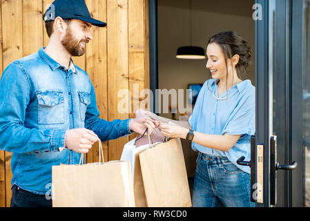 Uomo di consegna portando un po' di merci imballate in sacchi di carta per una giovane donna client a casa. Acquistare vestiti online e concetto di consegna Foto Stock