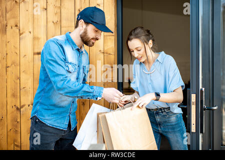 Uomo di consegna portando un po' di merci imballate in sacchi di carta per una giovane donna client a casa. Acquistare vestiti online e concetto di consegna Foto Stock