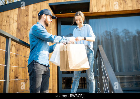 Uomo di consegna portando un po' di merci imballate in sacchi di carta per una giovane donna client a casa. Acquistare vestiti online e concetto di consegna Foto Stock