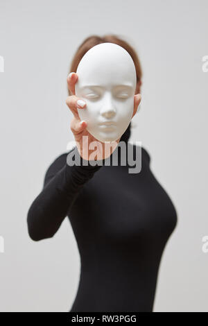 Giovane donna in un maglione nero con maschera in gesso in mano al posto della faccia su uno sfondo bianco. Concetto le maschere che indossiamo Foto Stock