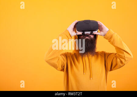 Hispter con barba lunga indossando felpa con cappuccio giallo e la realtà virtuale auricolare su sfondo giallo. Uomo bello. Barba lunga. Foto Stock