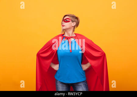 Giovane donna in costume superheros erge orgogliosamente su sfondo giallo. Super poteri. Red cape. maschera rossa. Foto Stock