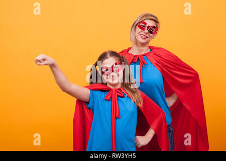 Bambina vestito come superheros tenendo il suo pugno in aria con sua madre su sfondo eyellow. Madre e figlia con red cape e maks. Foto Stock