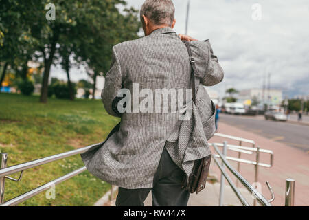 Un capelli grigi uomo anziano in una giacca grigia con un ombrello sotto il suo braccio e una pelle marrone a sacchetto sulla sua spalla correndo giù per la strada. Vista posteriore Foto Stock