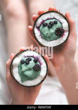 Due metà di noce di cocco con il frullato di yogurt o nelle mani di una donna. Blue spirulina smoothie in noce di cocco con frutti di bosco surgelati. Colazione sana nozione. Idee e ricette per una sana colazione vegan. Spazio di copia Foto Stock