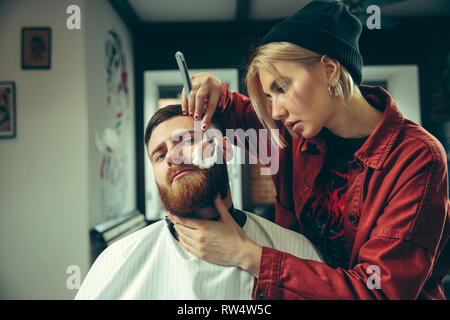 Client durante la rasatura della barba in barberia. Barbiere femmina al salone. La parità tra i sessi. Donna nel maschio professione. Foto Stock