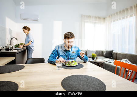 L'uomo mangiare insalata sana nel salotto del moderno appartamento con donna lavaggio piatti sullo sfondo Foto Stock