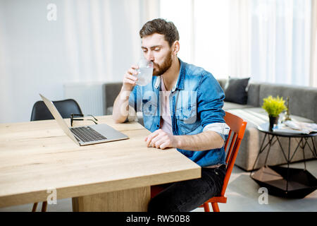 Uomo di bere medicinali dal mal di testa mentre si lavora con il portatile a casa Foto Stock