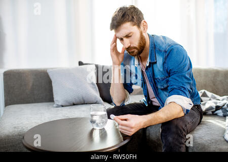 Uomo di bere alcuni farmaci sensazione di cattivo o aventi i postumi della sbornia dopo il partito di alcool, comodamente seduti sul divano di casa Foto Stock