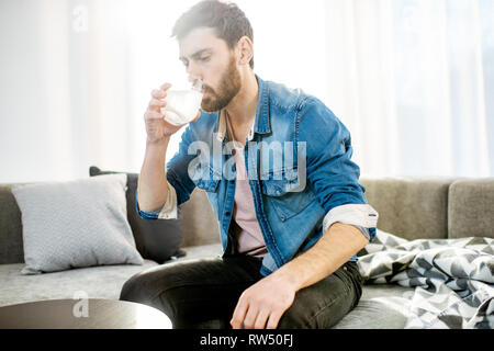 Uomo di bere alcuni farmaci sensazione di cattivo o aventi i postumi della sbornia dopo il partito di alcool, comodamente seduti sul divano di casa Foto Stock