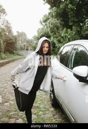 Giovane donna felice in sportswear allungamento della gamba vicino automobile nel giardino tra alberi verdi Foto Stock