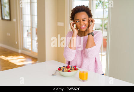 Giovane africano donna americana avente una sana prima colazione al mattino a casa che copre le orecchie con le dita con espressione di fastidio per il rumore di forte Foto Stock