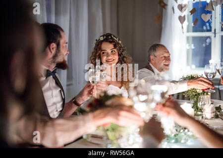 La sposa e lo sposo e gli altri ospiti seduti a un tavolo in un matrimonio, bicchieri tintinnanti. Foto Stock