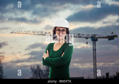 Bella fiducioso donna ingegnere sorridente positivo con gli occhiali e casco di protezione, tenendo le braccia incrociate, su costruzioni di sfondo del sito wi Foto Stock